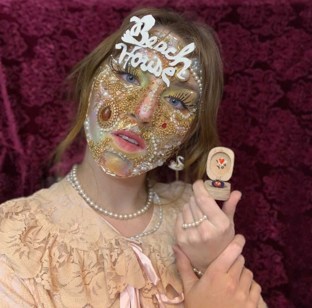Bergman wears layers of rainbow-colored makeup, very long black eyelashes, white and gold beads and gems, and a sign on her forehead that says "Beach House" in white. Her head is tilted; she's not smiling. One earring looks like a swan. She wears pearl jewelry and holds an open ring box. The inside of the lid of the ring box says "I [heart] You." 
