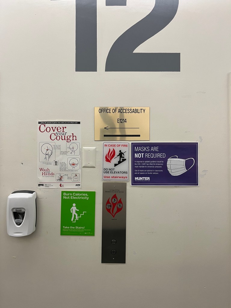 the wall between the elevators on the 12th floor of Hunter's East building, where the Office of Accessibility is. There are elevator buttons, a soap dispenser, a sign saying "cover your cough," a sign saying masks aren't required, a fire safety sign, a sign suggesting people take the stairs to burn calories and save electricity, and a sign pointing to the Office of Accessibility