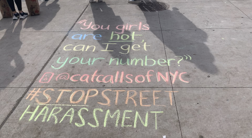 Colorful chalk on the sidewalk quotes a cat call. It says, "You girls are hot, can I get your number?" In pink and yellow chalk, there is the the Instagram handle @catcallsofnyc. In orange and yellow chalk it says "#STOPSTREETHARRASSMENT". 