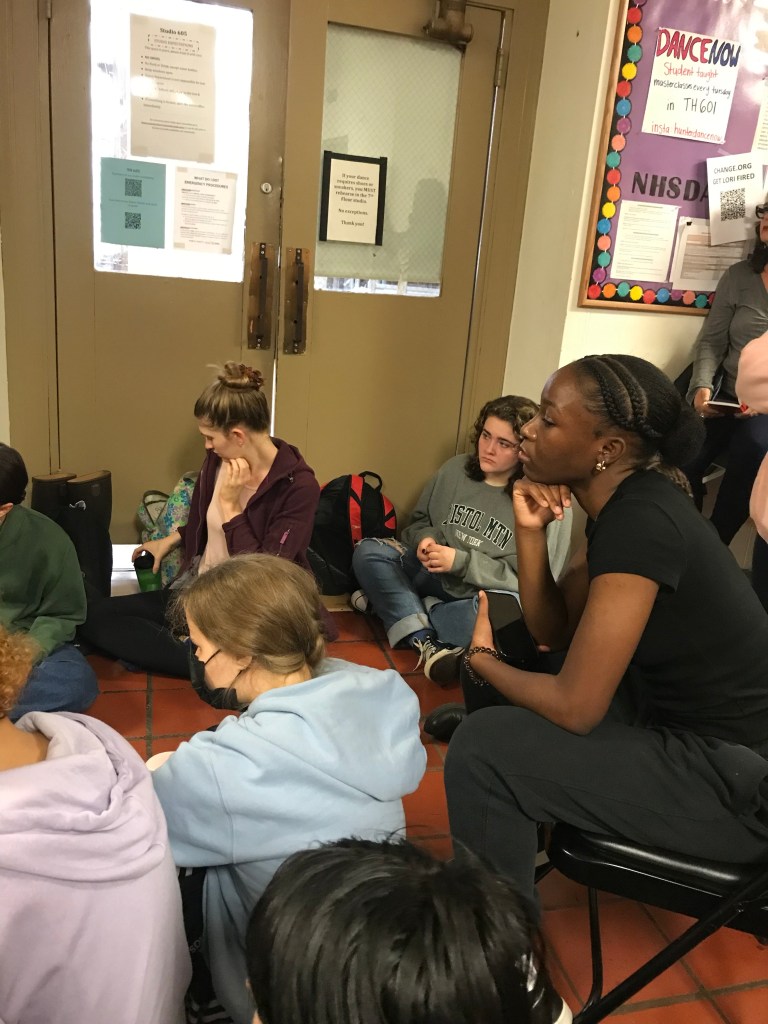 The image shows students sitting in the hallway. One student sits on a chair with their chin propped on their hand. Other students sit on the floor. All are looking toward the speaker at the center of the room. 