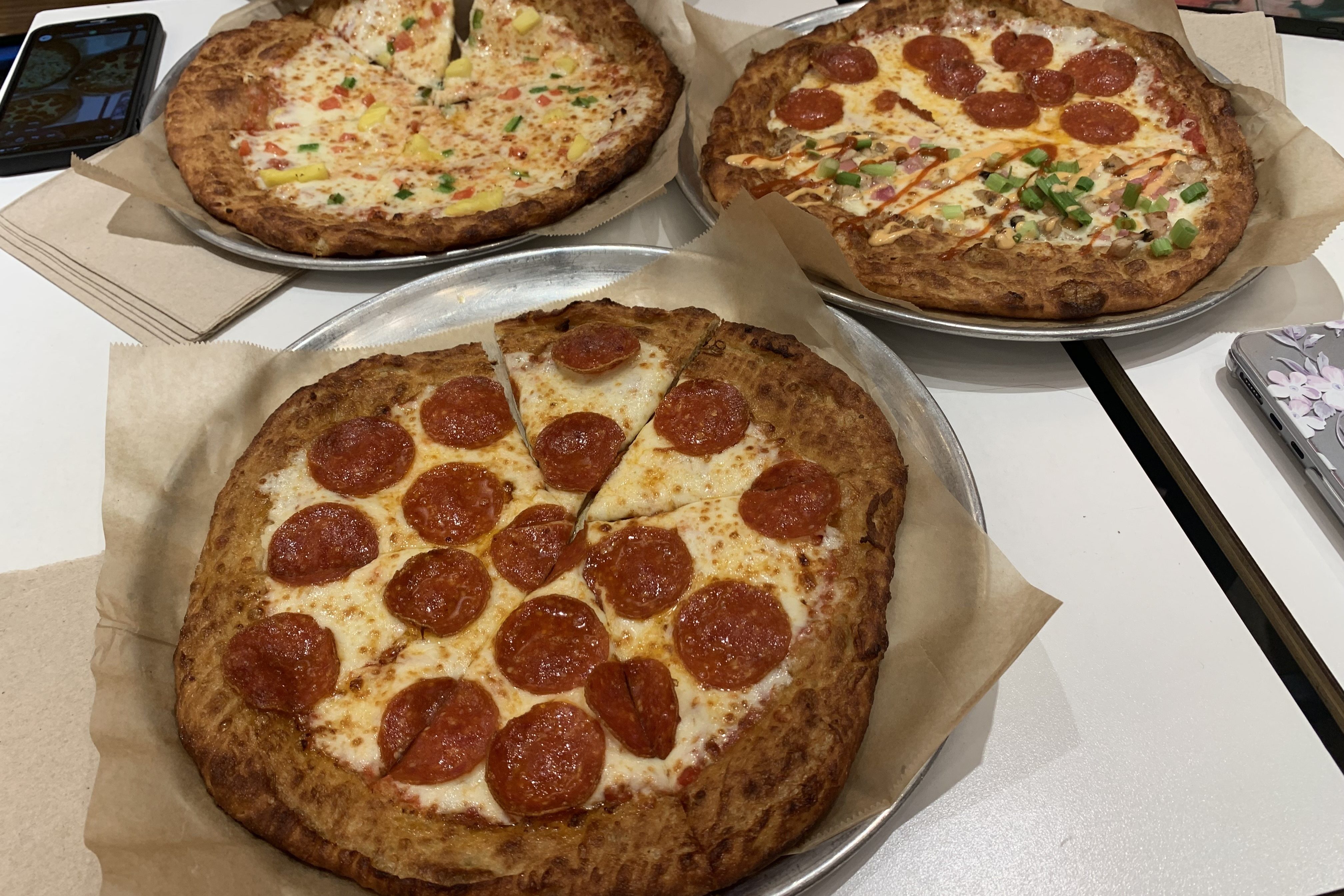 Photo displays three specialty pizza pies from Oath Pizza. One pie is covered in large slices of pepperoni. Another pie is half pepperoni and the other side has vegetables and sauces on top. Another pie is covered in veggies. All have a thick crust.